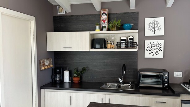 A modern kitchen with a dark countertop, light wood cabinets, a built-in oven, and a hanging shelf with decorative items. - Botanical Study 1 (San Carlos de Bariloche)