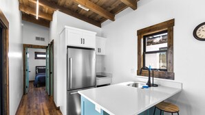 Cocina compacta con armarios blancos, refrigerador de acero inoxidable, un lavamanos y una ventana con un reloj.