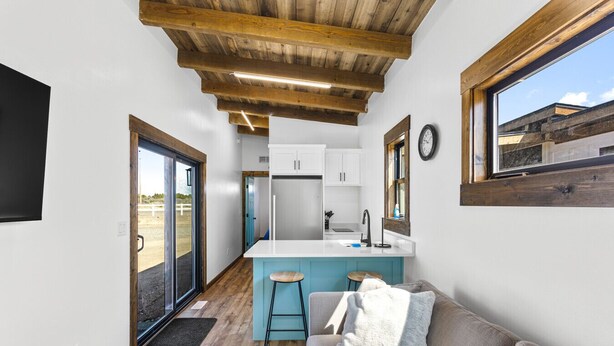 Un pasillo estrecho que conduce a una cocina con una isla central, un sofá con un cojín y un reloj en la pared.
