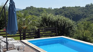Uma piscina retangular com borda de pedra, rodeada por espreguiçadeiras brancas e um guarda-sol azul.