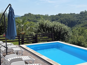 Une piscine rectangulaire bordée de pierres, entourée de chaises longues blanches et d’un parasol bleu.