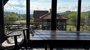 Um deck de madeira com guarda-corpos de metal, uma mesa e cadeiras, com vista para uma paisagem rural com um edifício de madeira e vegetação.