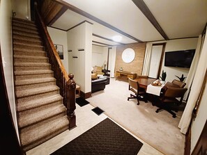 A staircase with wooden balusters, a dining area with a round table and chairs, a television mounted on the wall, and a brick accent wall. - This Old House / Cozy 3 BR near Sayers Lake (Howard)