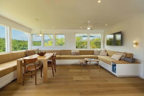 Ein modernes Wohnzimmer mit einer eingebauten Couch, Esstisch und Stühlen, großen Fenstern mit Blick und einem Deckenventilator.