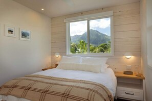 A bedroom with a bed, a nightstand, a bedside table, a window with a view of a mountain, and wall-mounted lights. - Hanalei River Cottages - Cottage Two, Luxury Private, Kauai North Shore stay (Hanalei)