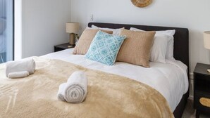 Een net opgemaakt bed met een beige deken en twee opgerolde handdoeken, een gestreept kussen en een effen kussen op een donker hoofdbord.