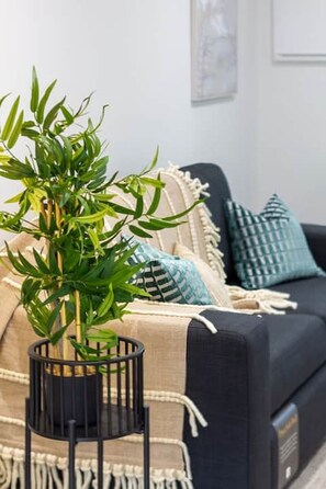 Ein Wohnzimmer mit einem schwarzen Sofa, blauen und weißen Kissen mit Muster, einem schwarzen Metall-Pflanzentisch und einer beigefarbenen Decke.