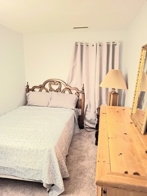A bedroom with a wooden bed, a bedside table with a lamp, a mirror, and a window with curtains.
