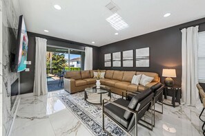 A modern living room with a sectional sofa, a glass coffee table, and a view to the outside. - Air Hockey | Classic Arcade | Fireplace | Resort Pool | Mins to Disney World: Storybook Villa (Kissimmee)