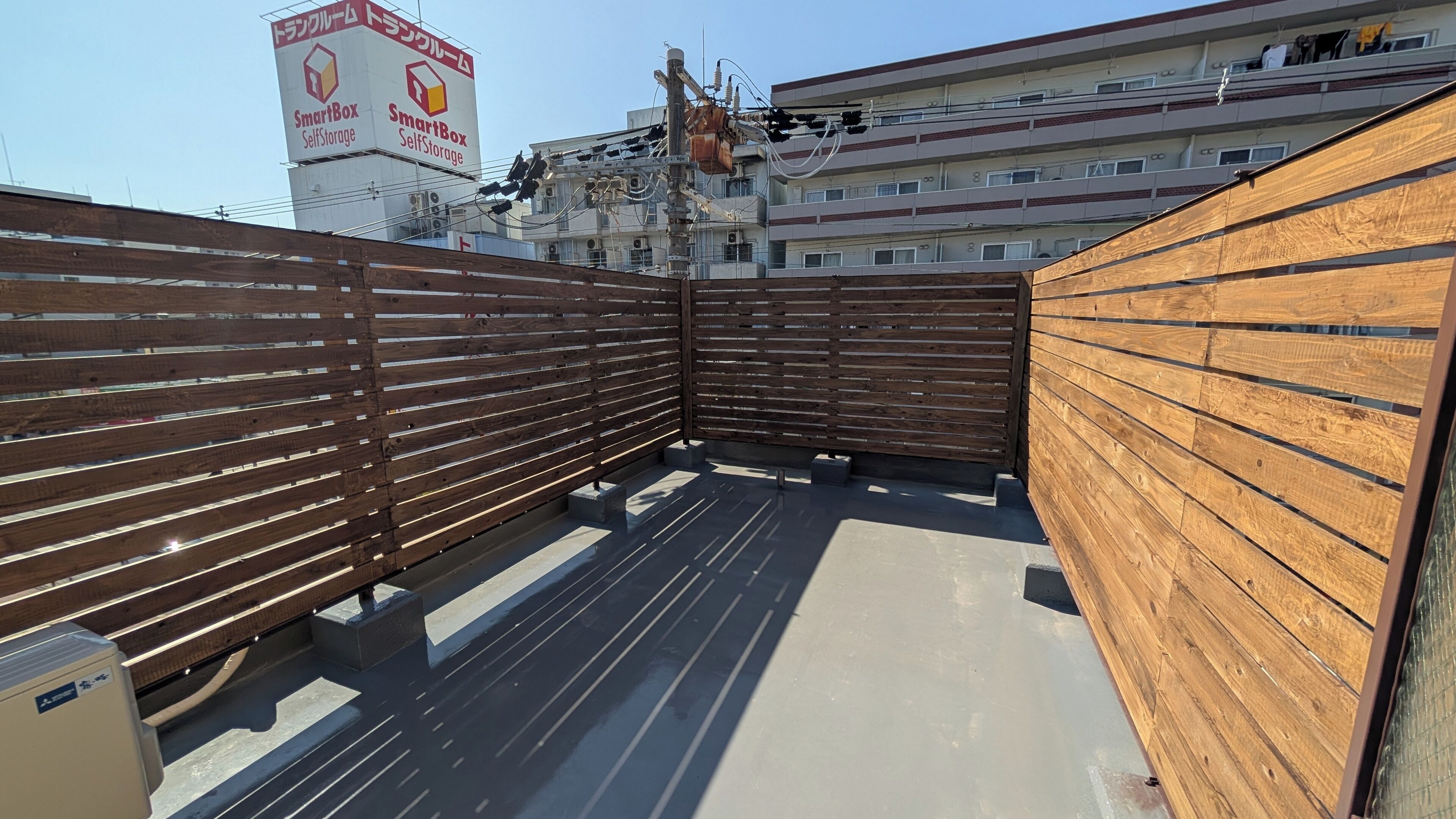 Una terraza en la azotea con cerramiento de madera, suelo de hormigón y un edificio con el rótulo SmartBox al fondo.