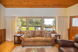 A living room with a brown leather sofa, two side tables with lamps, a wooden coffee table, and large windows with curtains. - Private Hilltop Cabin Near Yosemite, California (Mariposa)