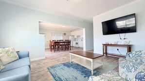A living room with a blue sofa, a patterned armchair, a coffee table, a TV mounted on the wall, and a kitchen in the background. - Beautiful 7BR Beach House, Pool, Walk to Beach (North Myrtle Beach)