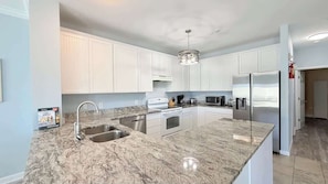 Una cocina moderna con armarios blancos, encimeras de granito, electrodomésticos de acero inoxidable y una lámpara colgante.