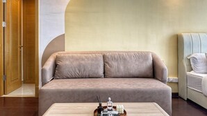 Una habitación de hotel moderna con un sofá color crema, una mesita con un set de amenities y una cama con ropa blanca.
