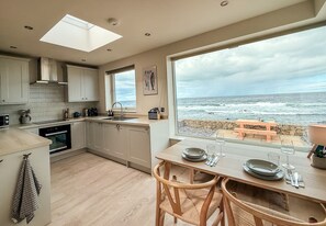 A modern kitchen with a large window overlooking the sea, wooden dining table set, and a view of the beach. - Writer s Retreat - Peaceful Cosy Seafront Retreat for Two (Fraserburgh)