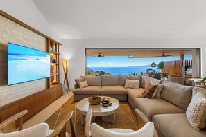 A modern living room with a large flat-screen TV, a sectional sofa, a coffee table, and a view of the sea. - Ocean-View Iyari Villa, Pool, Cook & Golf Cart (Corral del Risco)