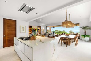 A modern kitchen with a central island, dining area with wooden chairs, and a large window overlooking a scenic view. - Ocean-View Iyari Villa, Pool, Cook & Golf Cart (Corral del Risco)
