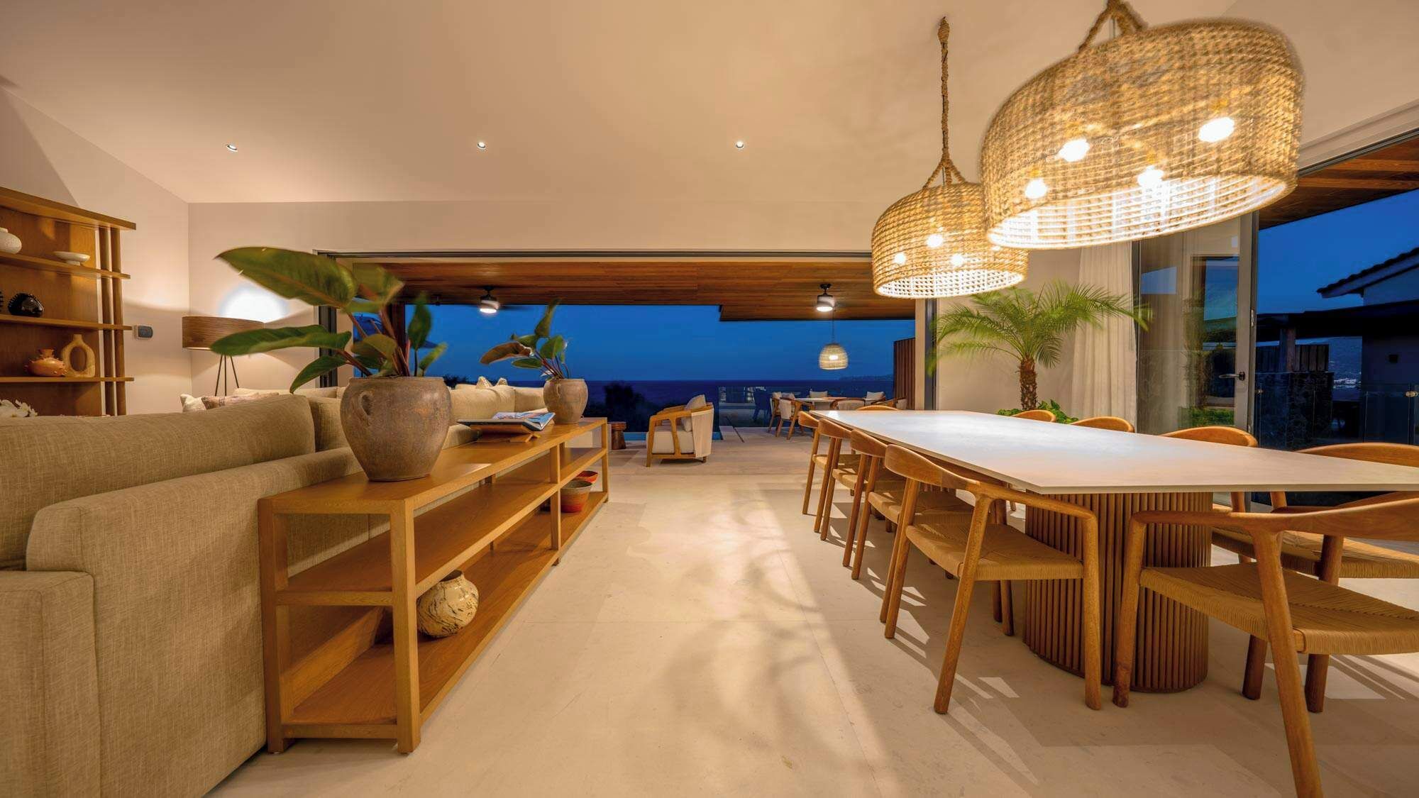 Una sala moderna con sofá, mesa de centro de madera y comedor con sillas. Hay plantas en macetas y vistas al exterior.