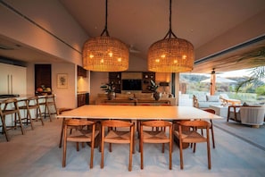 A modern dining area with a large table and chairs, a kitchen in the background, and a view of the outdoors. - Luxury Iyari Villa Inside Punta Mita Ocean View (Corral del Risco)