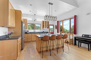 Una cocina moderna con armarios de madera, una isla central con taburetes altos, un piano y ventanas amplias con vistas a zonas verdes.