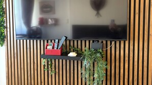 A flat-screen TV mounted on a wooden slat wall, a small shelf with remote controls, a plant, and a bench.