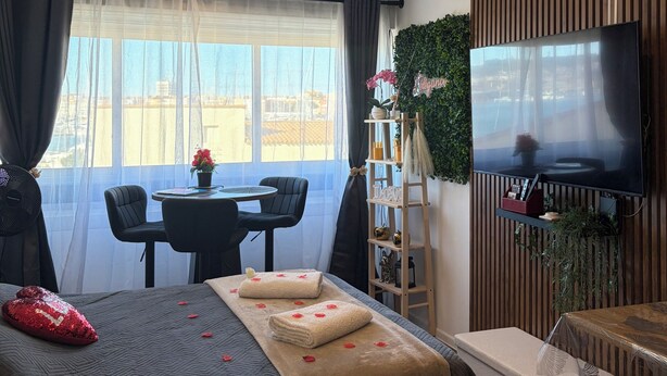 A hotel room with a bed, a TV, a dining table with chairs, a small balcony, and a view of the city.