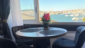 A round dining table with black chairs, a potted plant with red flowers, and a view of a marina with boats and buildings.