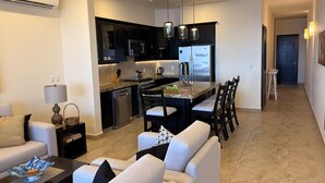 A modern living room with a sofa, coffee table, and a dining area with chairs and a kitchen in the background. - One Bedroom Oceanfront - Puerto Escondido 1423 (Bajos de Chila)