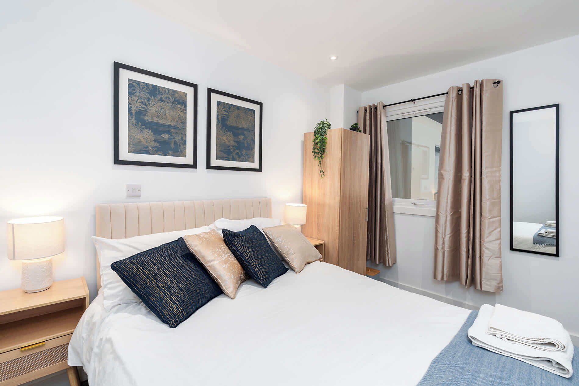 Una habitació d'hotel amb un llit, dues fotografies emmarcades, una finestra amb cortines, una làmpada de nit i un mirall.