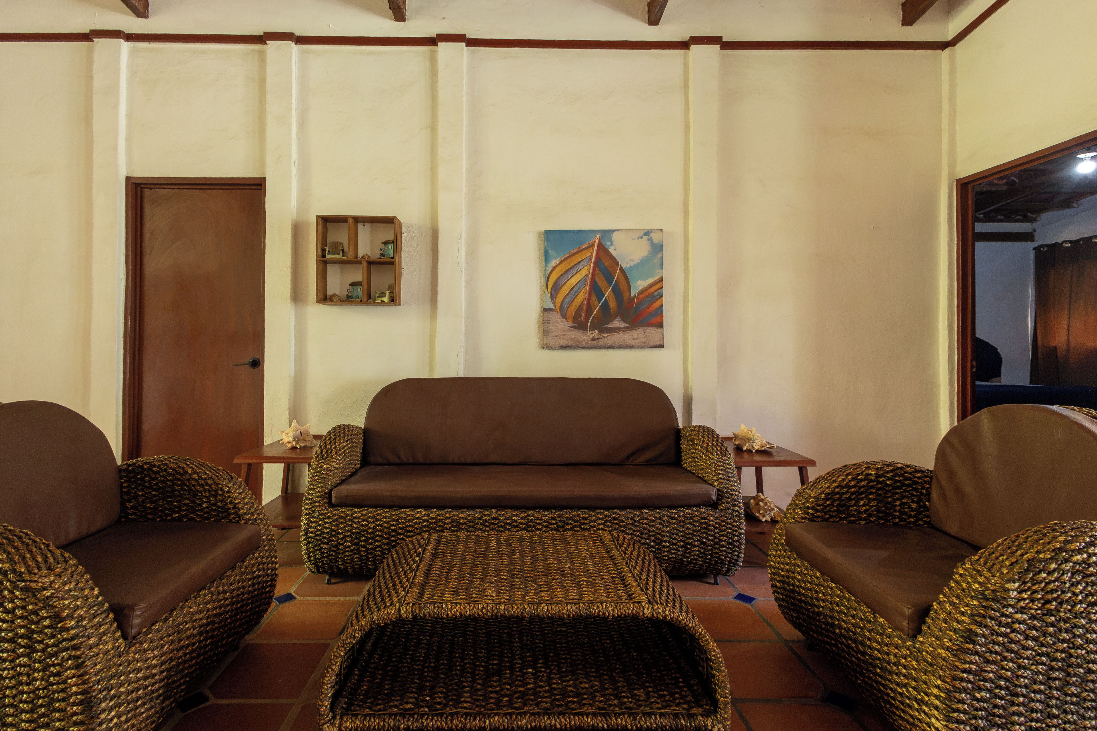Una sala de estar con un sofá marrón, sillones de mimbre, una mesita y un cuadro en la pared.