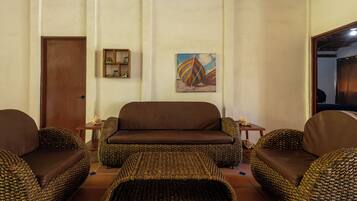 Una sala de estar con un sofá marrón, sillones de mimbre, una mesita y un cuadro en la pared.