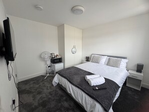 A hotel room with a bed, a bedside table, a nightstand, a wall-mounted TV, a fan, and a microwave. - Near Heathrow - Studio - WiFi - Parking - Sleeps 2 (Uxbridge)