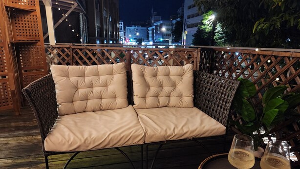 Un área de estar al aire libre con asientos acolchados, una valla de mimbre, una mesa redonda con dos vasos de una bebida transparente y vistas a un paisaje urbano nocturno.
