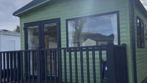 A small, single-story mobile home with a dark roof and light green siding, featuring large windows and a black railing.