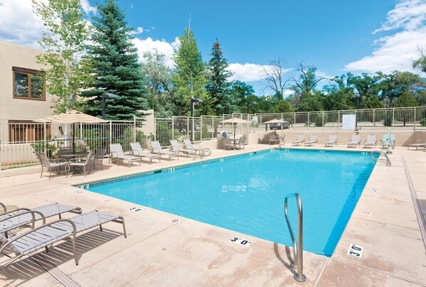 Una piscina rectangular con una escalera metálica, rodeada de sillones de descanso y sombrillas, con árboles y un edificio al fondo.