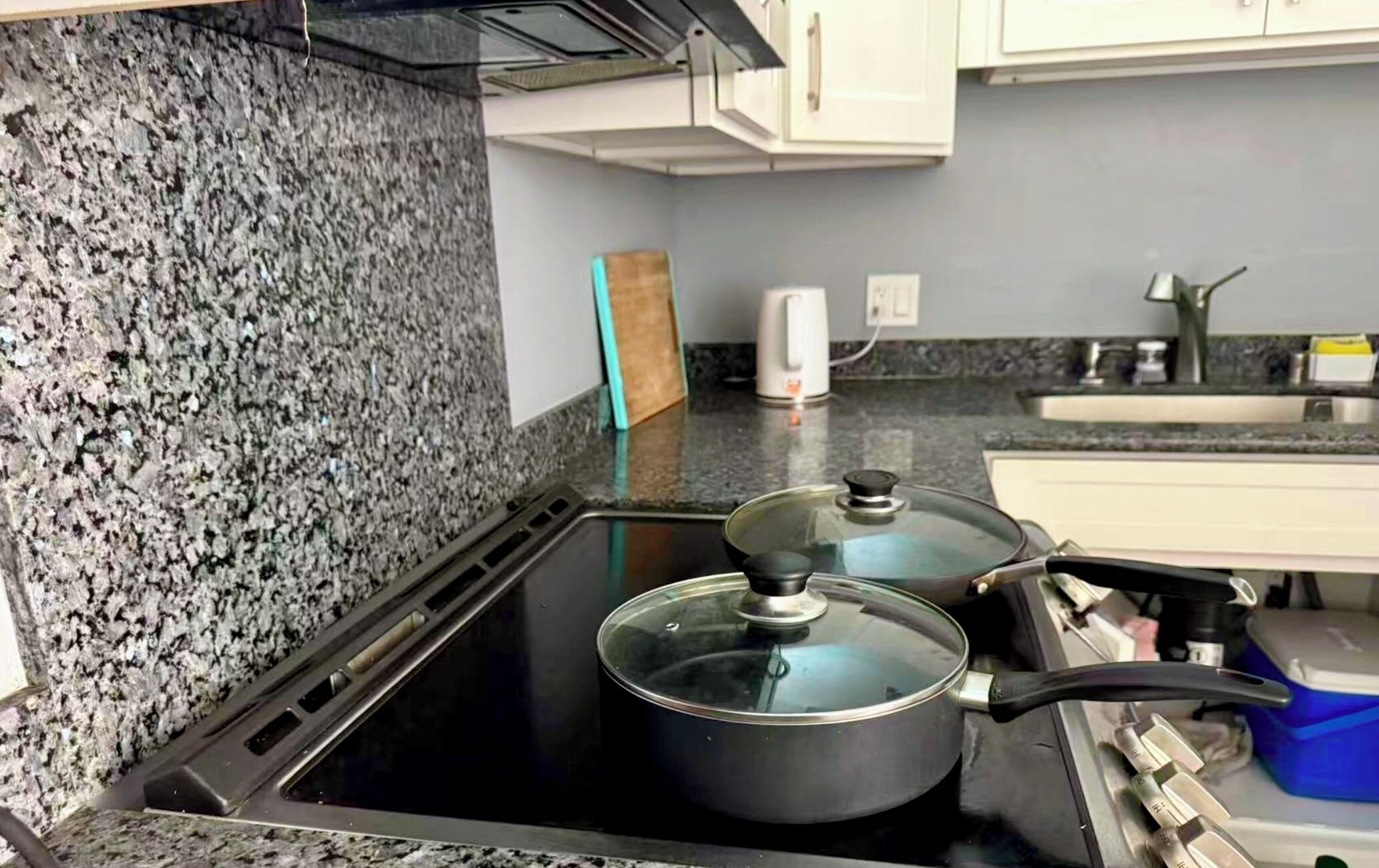 A kitchen with a granite countertop, a stove with pots, a coffee maker, and a blue storage box.