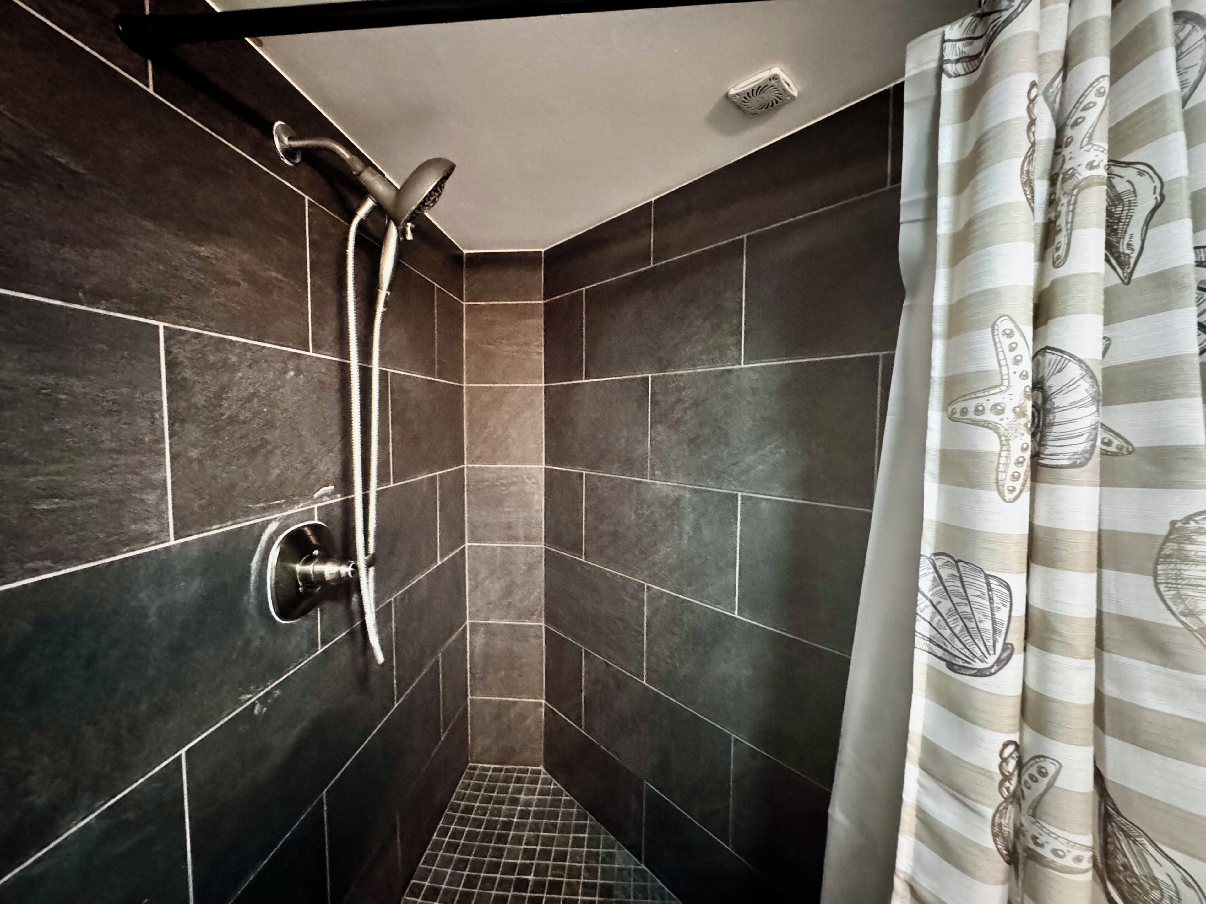Una ducha con cabezal de ducha de mano y llave de pared, paredes con azulejos y cortina a rayas.