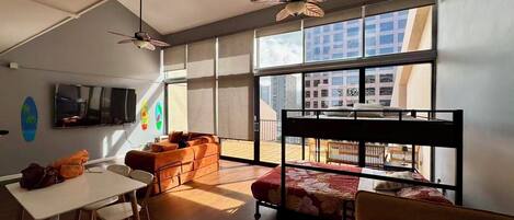 A modern room with a bunk bed, a dining table, and chairs. There is a TV mounted on the wall and a ceiling fan.