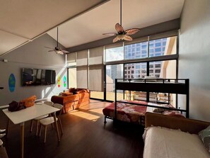 Ein modernes Zimmer mit einem Etagenbett, einem Esstisch und Stühlen. Ein Fernseher ist an der Wand montiert und ein Deckenventilator vorhanden.