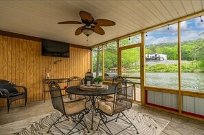 Eine überdachte Terrasse mit Tisch und Stühlen, einem Deckenventilator und Blick auf einen Fluss und Gebäude.