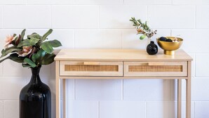 Pared de ladrillo blanco con un espejo redondo, una consola de madera con dos cajones, un jarrón negro y un cuenco dorado.