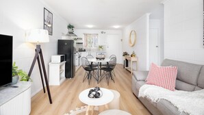 Una sala moderna con un televisor de pantalla plana, un comedor con mesa y sillas, y un sofá gris con un cojín rojo.
