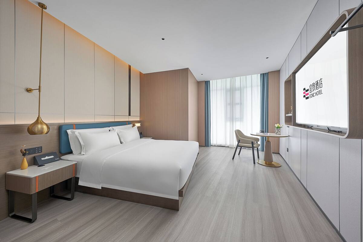 Habitación de hotel con cama, mesita de noche, lámpara, silla y televisión mostrando la marca del hotel.