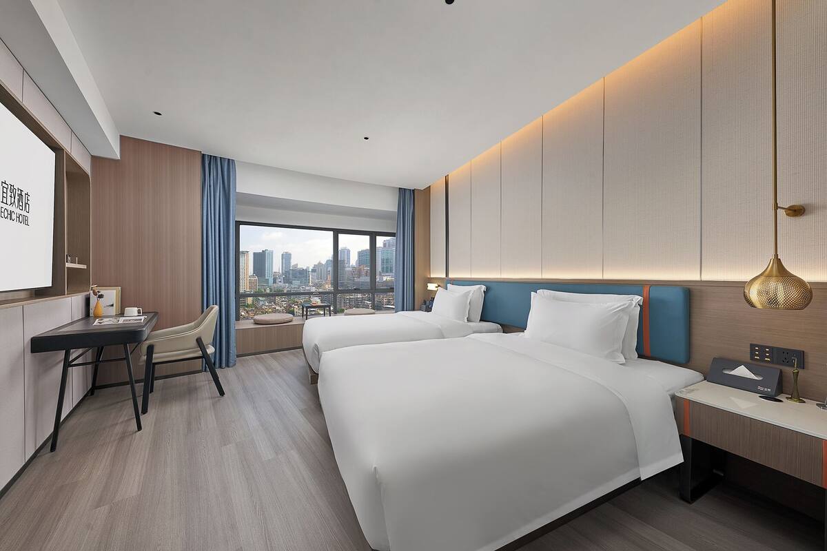 Habitación de hotel con dos camas, un escritorio, una silla, un ventanal grande con vista a la ciudad y una lámpara moderna.