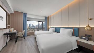 A hotel room with two beds, a desk, a chair, a large window with a city view, and a modern lamp.