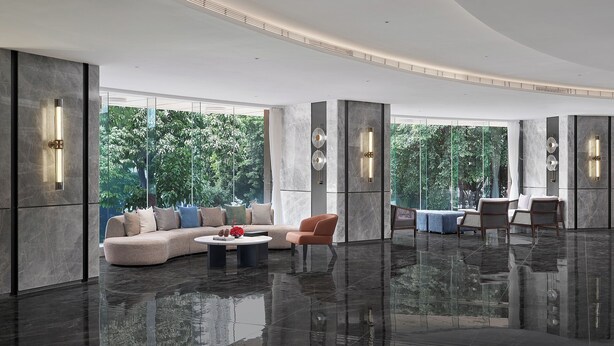 A spacious living area with a large sofa, a coffee table, and a chair. There are large windows providing a view of greenery outside.