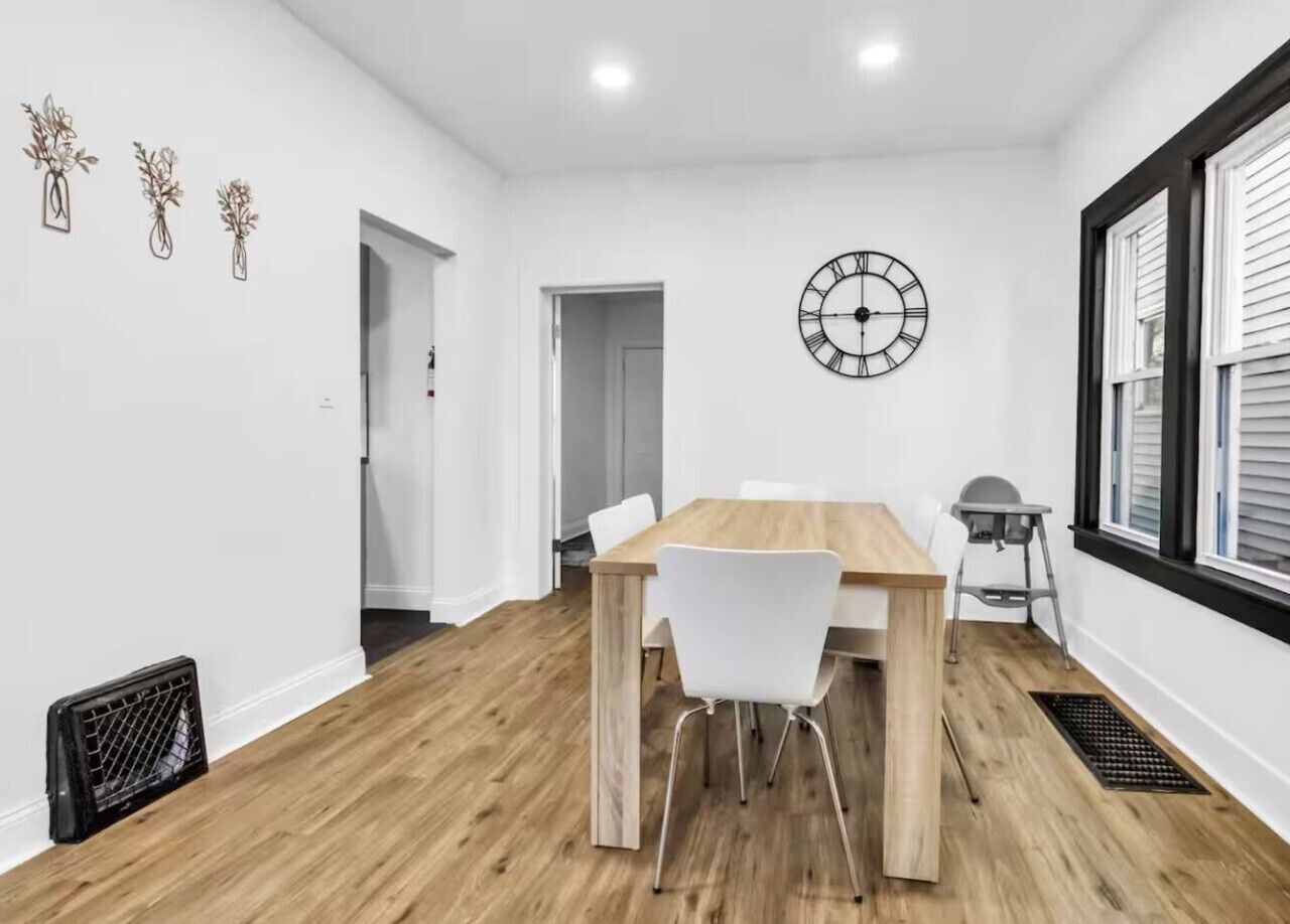 A modern dining room with a wooden table and white chairs, a wall clock, and a window with blinds.