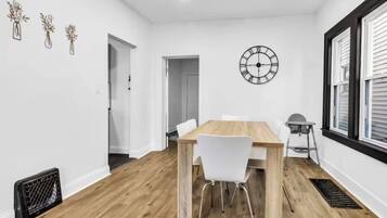 A modern dining room with a wooden table and white chairs, a wall clock, and a window with blinds.
