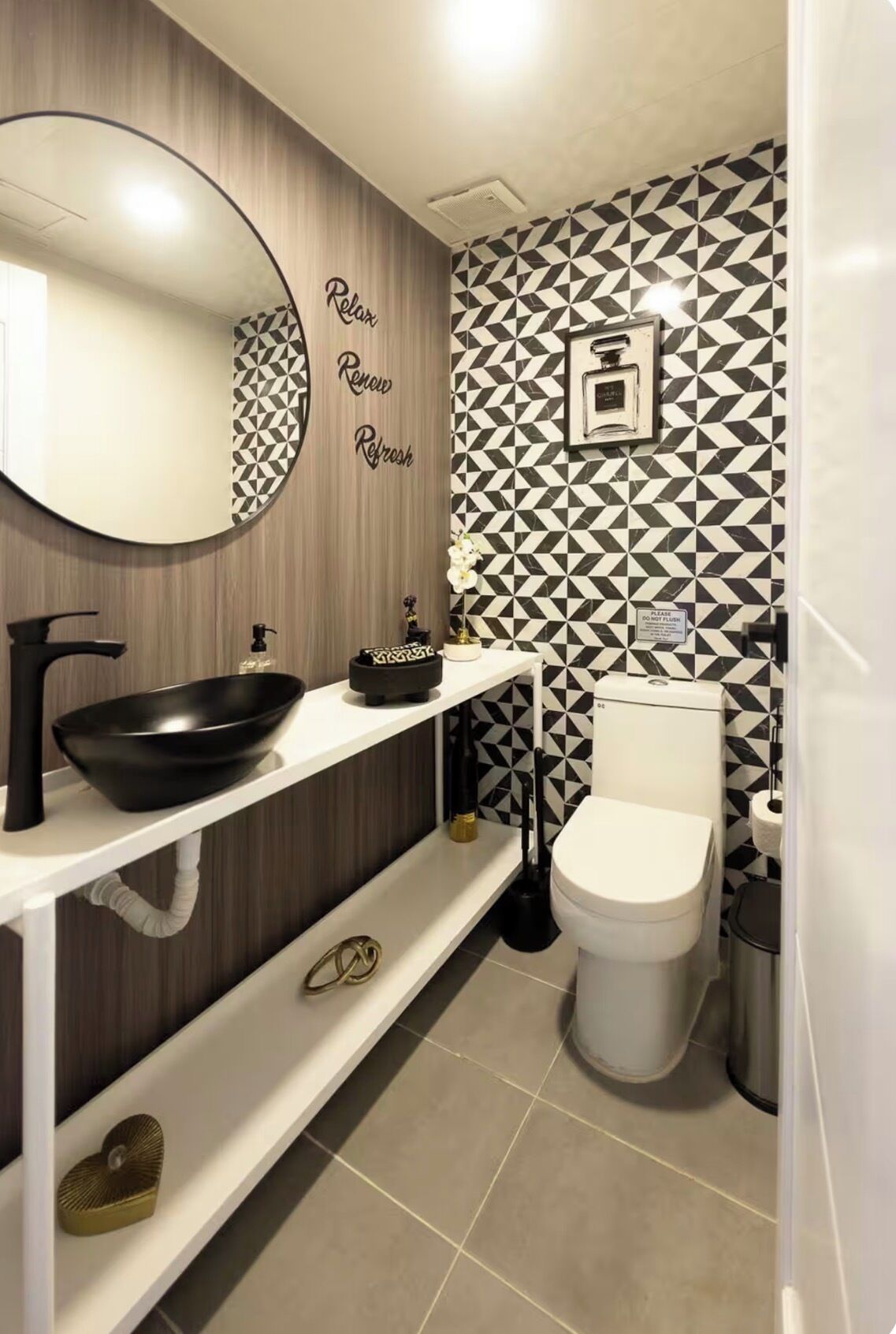 Une salle de bain moderne avec un mur recouvert de carreaux géométriques noirs et blancs, un miroir rond orné d'une inscription « Relax », un lavabo noir et des toilettes.