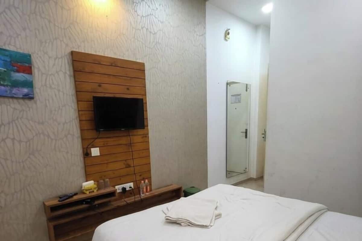 A bedroom with a bed, a TV mounted on a wooden panel, a small shelf with toiletries, and a door leading to a bathroom.